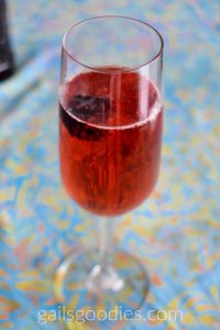 A champagne flute fille with cherry red liquid. bubbles line the top and there is a single blackberry floating in th eclass on the left side.