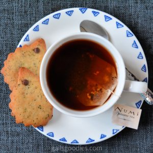 A tea cup and saucer viewed from above. The cup is filled with tea and the top of the tea bag is just visible in the liquid. The tag on the teabag says "Numi organic tea." There is a spoon on the saucer to the right of the cup and two small golden brown hedgehog shaped cookies to the left of the cup. The hedgehog cookies are golden brown and flecked with dark brown specks. Each cookie has a mini chocolate chip eye.