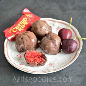 Three Cherry Ripe Truffles are arranged in a triangle on a plate. There are two cherries next to the truffle on the right. There is a small cherry ripe bar in its wrapper behind the truffles on the left. A partially eaten truffle is in front of the three truffles revelaing the red cherry coconut insides.