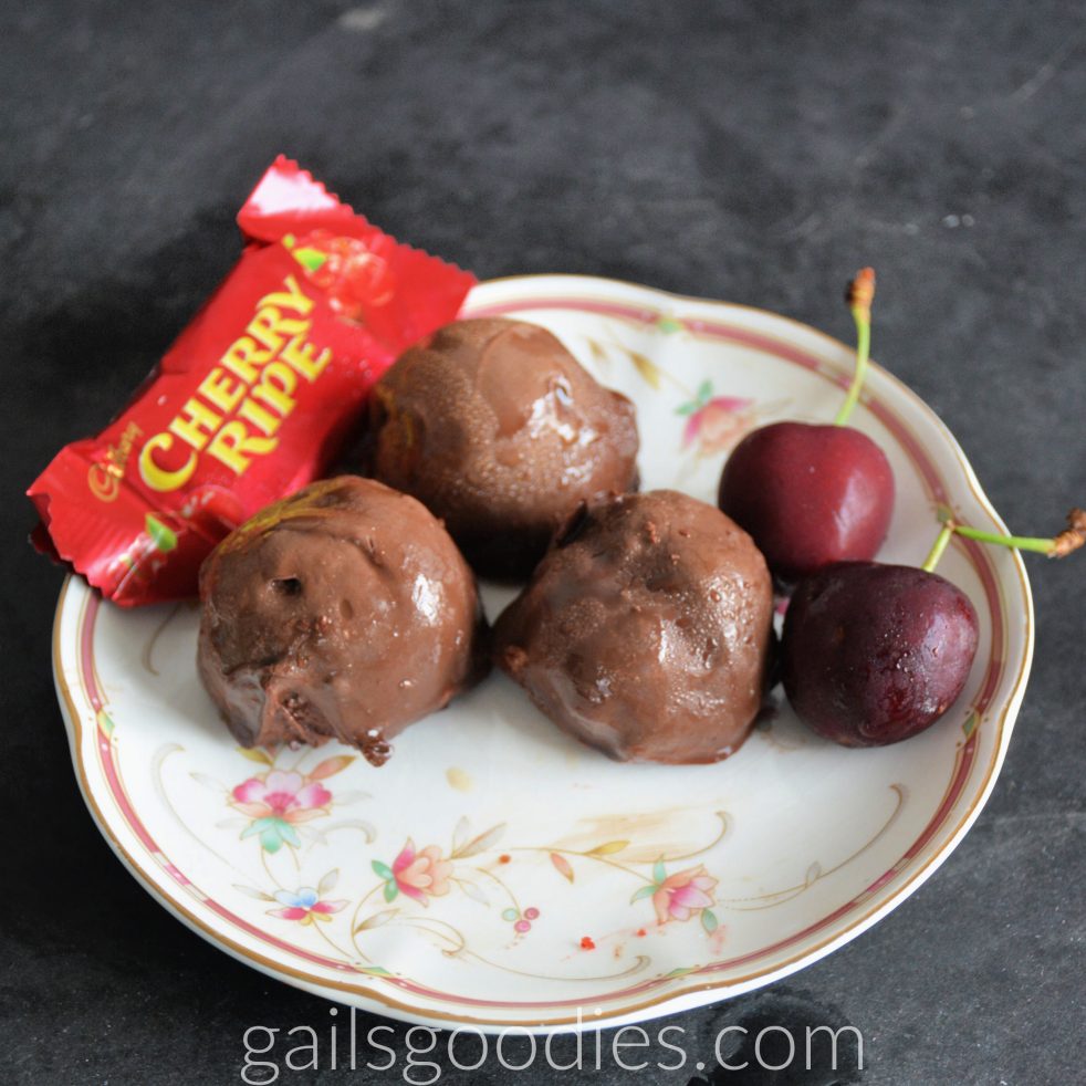 Three Cherry Ripe Truffles are arranged in a triangle on a plate. There are two cherries next to the truffle on the right. There is a small cherry ripe bar in its wrapper behind the truffles on the left.