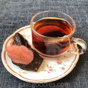 Two chocolate cherry madeleines are on a plate to the left of a clear glass cup of tea. The madeleine in front is pink on top and dark broen on the bottom. Only the dark brown top of the madeleine in the back is visible.