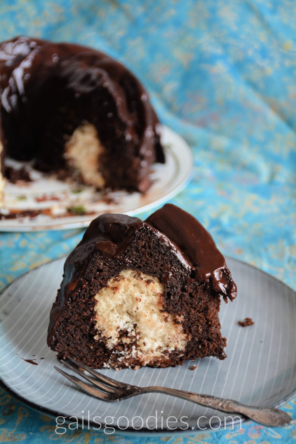 A slice of chocolate fudge macaroon bundt cake stits on a grey plate with white strips. cream colored macaroon filling is surroundd by moist, dark chocolate cake. The slice is covered with a generous amount of fark chocolate ganache. In the background on the left side of the photo, the rest of the cake sits on a white plate.