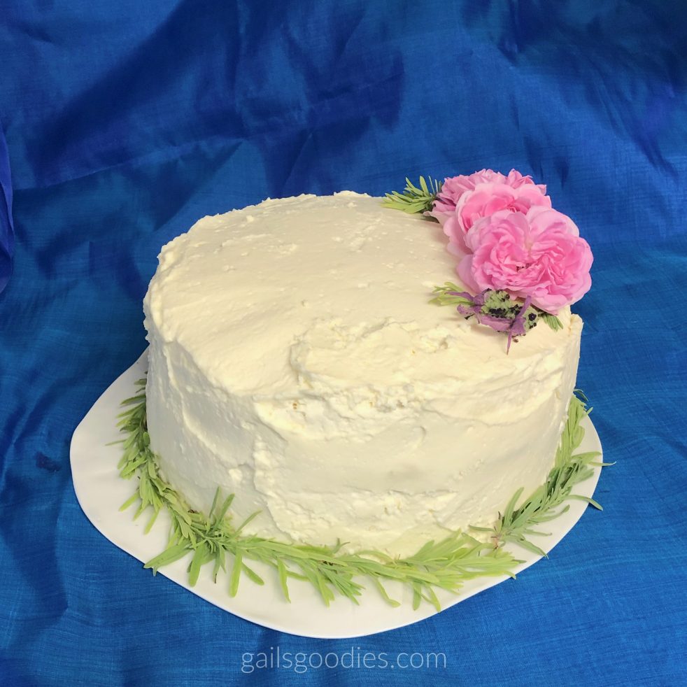 A whole cake sits on a white plate with a vibrant blue background. The cake is about 6 inches tall and is covered with creamy white frosting. There is a ring of light green lavender leaves around the base of the cake. Three pink roses, two lavender flowers and a few lavender leaves are arranged in an arc on the top of the cake on right side.