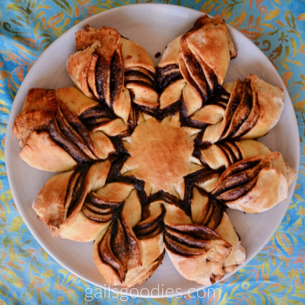 A loaf of mexican chocolate star bread viewed from above. I the center is an 8 pointed star.The golden bread star is surroundd by about 1 inch of chocolate filling. A brange of bread goes out from each point of the star. The left side of each brange shows 4 layers of bread with chocolate and cinnamon in between. The right side of each branch also has 4 layers. The top left side of each branch is tucked under the right side. The pattern looks like vees going from each point of the star.