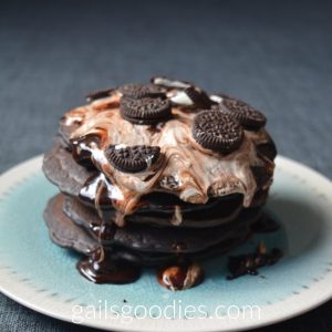 A stack of black chocolate oreo pancakes topped with dark chocolate fudge sauce and marshmallow cream. Broken mini oreos are scattered across the top.