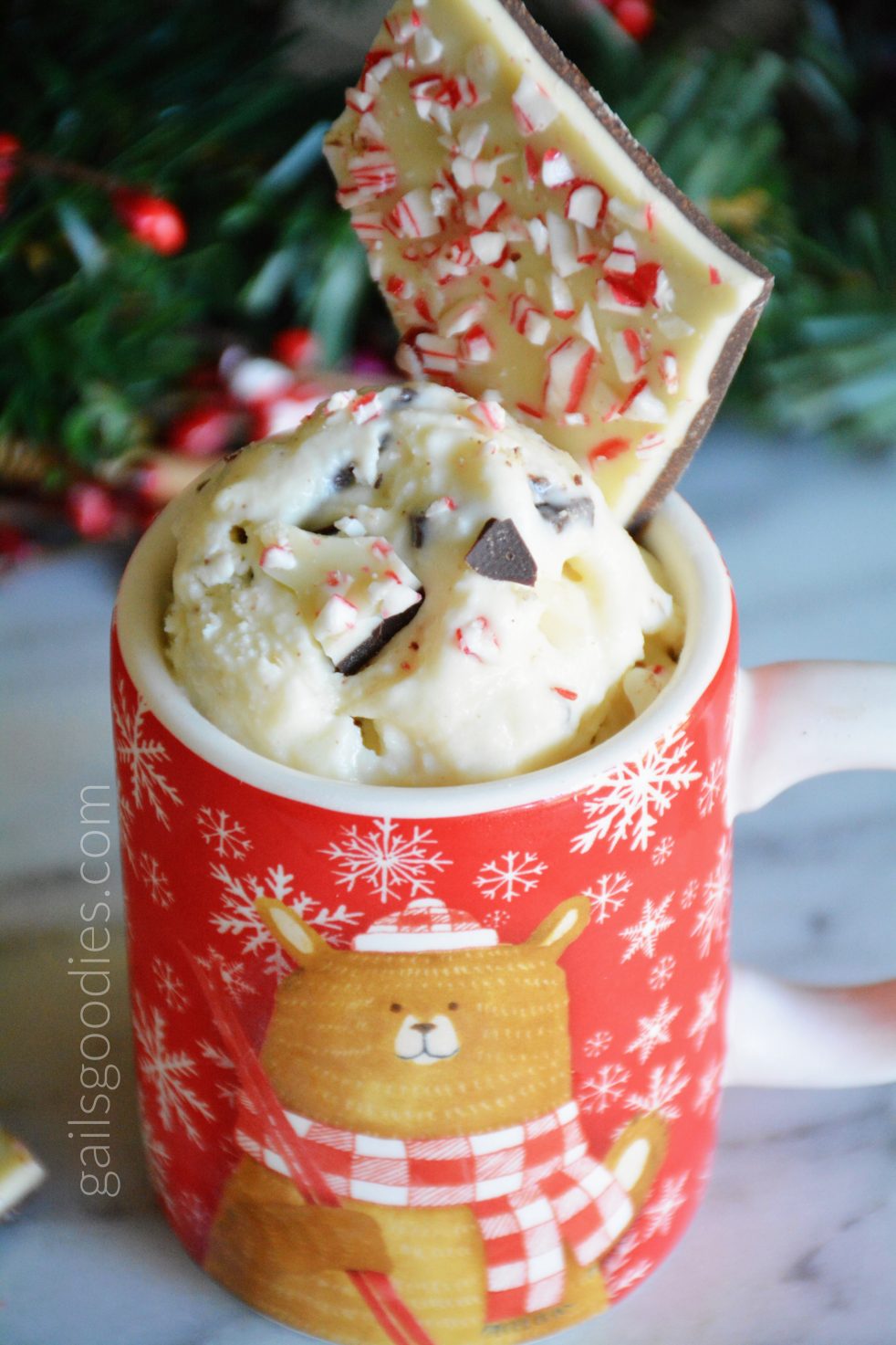 A red coffee mug with a bear and snowflakes is filled with peppermint bark ice-cream. A scoop of ice-crean extends above the top of the mug. Creamy white ice-cream is flecked with bits of peppermint, dark chocolate and peppermint bark. A small piece of peppermint bark garnishes the ice-cream at the back of the mug.