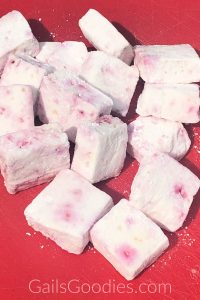 Peppermint stick marshmallows on a red board. The square marshmallows are scattered from the upper left corner to the lower right corner. The marshmallows are slightly pink with dark pink dots.