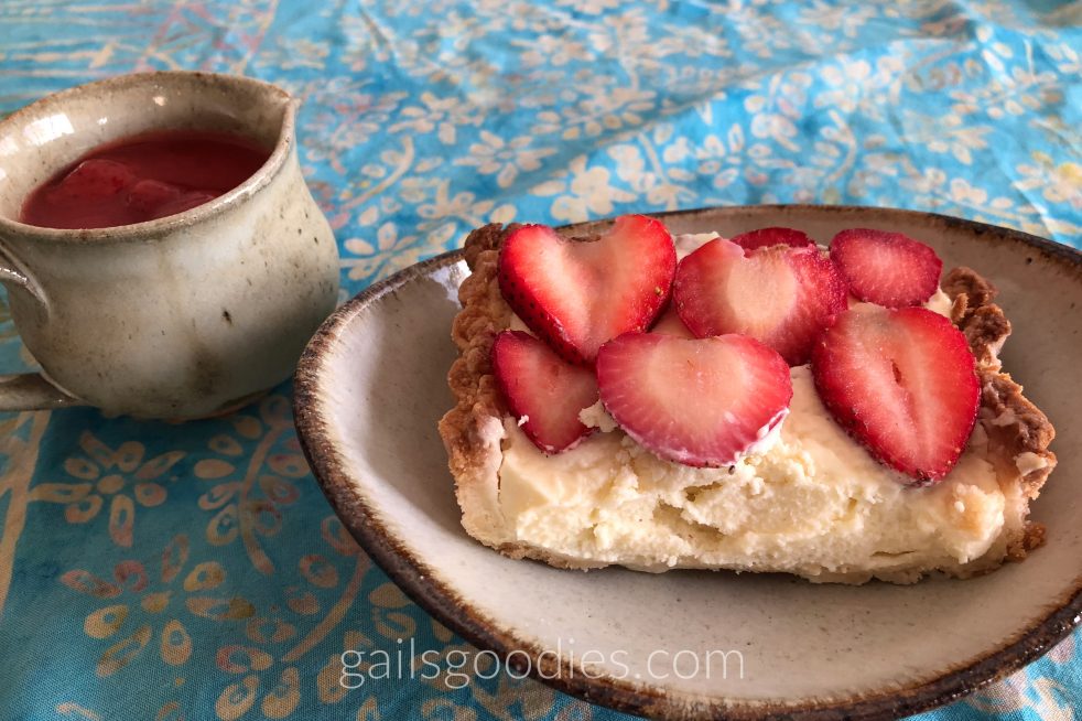 A slice of strawberry mascarpone tart on a white plate with brown edges. The cut side of the tart faces the front of the photo revealing about an inch of mascarpone filling topped with sliced strawberries. A small ceramic pitcher filled with strawberry sauce sits behind the plate to the left.
