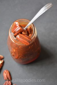 This is a slightly offset view of the top of a wide-mouthed glass bottle of bourbon caramel pecan sauce. The handle of a silver spoon extends outward from the top of the bottle towards the upper left corner of the photo. The bowl of the spoon is inside the top of the jar and is holding up four of the caramel covered pecans. One of the caramel covered pecans hangs over the bottomm edge of the jar mouth. Another pecan is slightly elevated above the top of the jar opening. There are three pecans scattered in the lower left corner of the photo.
