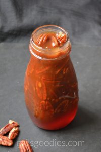 This is a side view of a clear glass wide-mouthed bottle filled with bourbon caramel pecan sauce. The view is from about a thirty degree angle above the bottle so the top is also visibe. The medium brown caramel coats the three pecans in the opening at the top of the bottle. Pecans also line the sides of the bottle almost down to the bottom. The bottom eighth of the bottle is clear golden honey colored caramel. There are four pecans scattered in the bottom left corner of the photo.