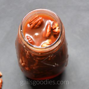 An open glass bottle of Bourbon Caramel Pecan Sauce is viewed from the top and at a slight angle to the front. Five pecans are peeking out from the thick caramel sauce in the wide opening of the bottle. 