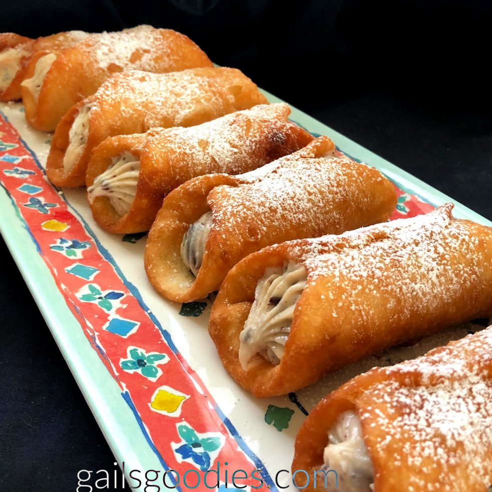 Six chocolate cherry cannolis are arrange side by side on a rectangular tray. The tray is teal and has a red-orange band around the edge. The band has alternating blue and yellow decorations. The tray is angled diagonally across the photo the the ends of the cannolis are visible as well as the sides and top. The golden brown fried shells are filld with a creamy white filling dotted with small brown and deep red flecks. The cannolis are dusted generously with powdered sugar.