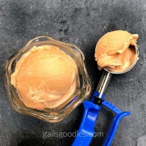 This is a view from the top of a glass ice-cream bowl filled with a single scoop of no churn caramel ice-cream. The deep golden brown icecram is slightly melted on the sides. To the right of the bowl there is a blue-handled ice-cream scoop with a scoop of caramel ice-cream still in the bowl. The bowl of ice-cream and the ice-cream scoop are sitting on a slate surface.