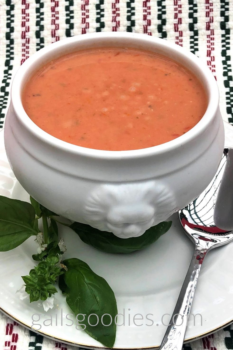 A white footed bowl filled with tomato basil soup sits on a white plate with a gold rim. There is a spoon on the plate to the right of the bowl and a sprig of basil on the plate to the left of the bowl. The front of the bowl is decorated with a lion's head. The tomato basil soup is a medium orange red and is speckled with dark green flecks. The view is from about 45 degrees above the bowl.