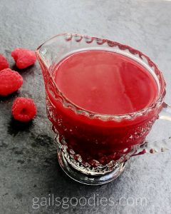 This is a view from about 45 degrees above of a cut glass pitcher filled with Grand Marnier Raspberry Sauce. The bright red cause contrasts with the grey slate table. There are three raspberries scattered on the table to the upper left of the pitcher.