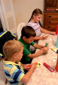 Three kids sitting at a table decorating cookies. The view is from the side and above. They are facing the right side of the photo.