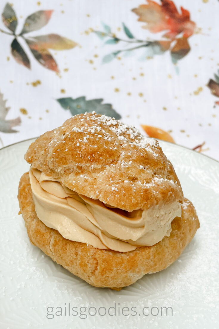 A single caramel apple cream puff sits on a white plate. The pastry shell is cut so the piped whipped cream is visible in a thick layer between the base and the top. The golden brown shell is dusted with powdered sugar