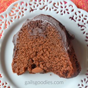 A slice of chocolate fudge bundt cake sits on a white plate with lacey edges. There is a circle of dark fudge surrounded by chocolate cake at the top of the slice. The thick dark chocolate ganache tops the cake and continues about two thirds down the sides.