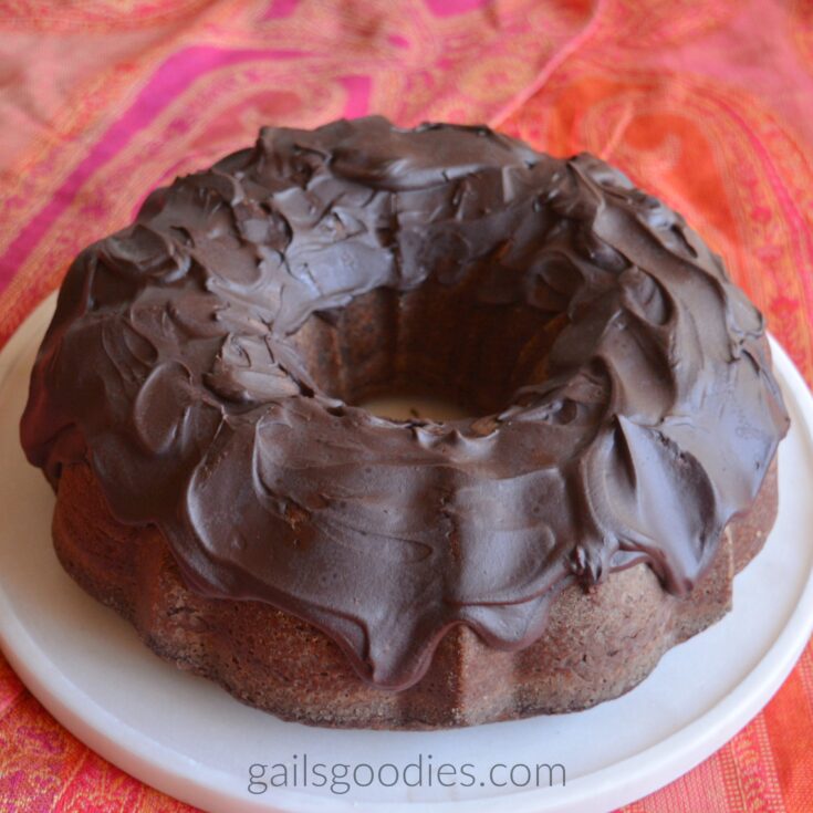 This is a view of the unsliced cake from about 45 degrees above. The top and about two thirds of the sides are covered in dark chocolate ganache. The cake sites on a white plate which is on a bright orange and pink cloth.