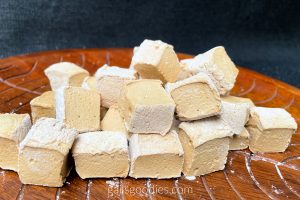 A small pile of espresso marshmallows are viewed close up on a wooden platter. There are two layers of marshmallows in the pile and the marshmallows are haphazardly arranged. The latte-colored marshmallows are cube-shaped and the tops are dusted with powdered sugar. The cut sides face th viewer