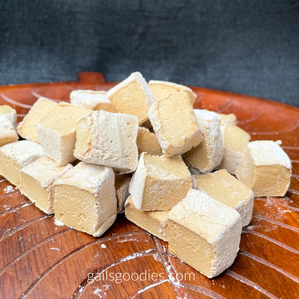A small pile of espresso marshmallows are viewed close up on a wooden platter. The latte-colored marshmallows are cube-shaped and dusted with powdered sugar.