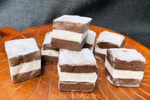 Six cube-shaped oreo marshmallows are scattered on a wooden platter. A seventh marshmallow sits on top of three marshmallows in the center. Each marshmallow has a dark chocolate base and top layer with a vanilla marshmallow layer sandwiched in between. The tops of the marshmallows are dusted with powdered sugar.